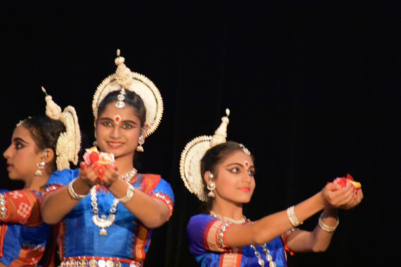Three Odissi dancers in traditional attire offering flowers on a decorated stage