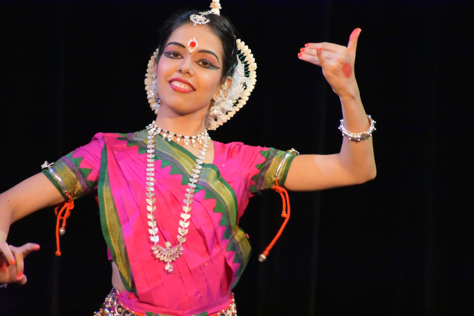 Odissi dancer in vibrant traditional attire with one hand elegantly raised, adorned with intricate jewelry