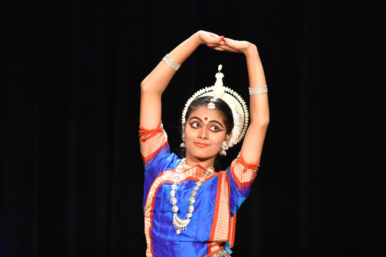 Odissi dancer in traditional attire with hands raised gracefully, adorned with intricate jewelry.