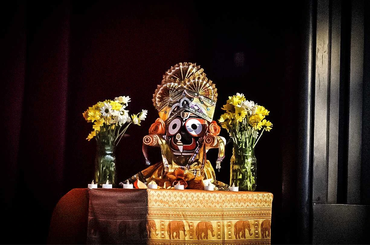 A decorated idol of the Hindu deity Jagannath on an altar, flanked by vases of flowers and candles against a dark background.