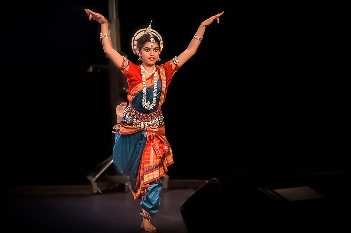 An Odissi dancer poised gracefully with arms raised against a dark stage background