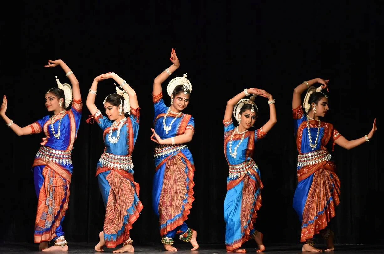 Five Odissi dancers in vibrant blue and maroon costumes performing synchronized poses with graceful hand and body movements on stage