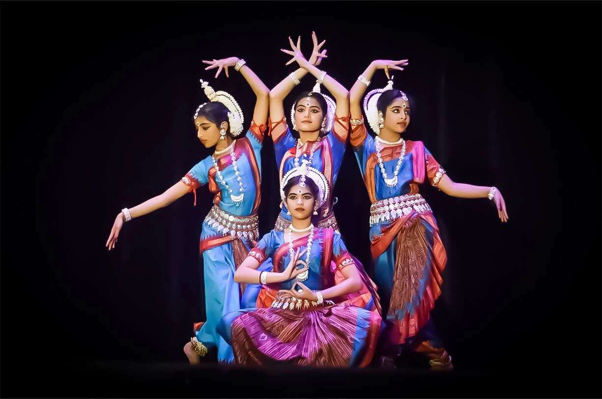 Four Odissi dancers in blue and maroon traditional costumes performing a symmetrical pose on stage with intricate hand gestures