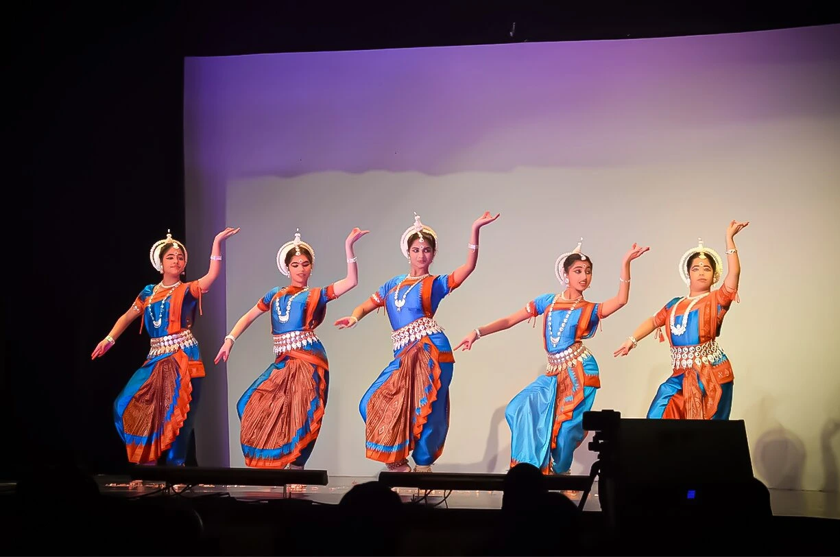 Group of Odissi dancers in blue and orange traditional costumes performing in graceful unison on stage with expressive poses