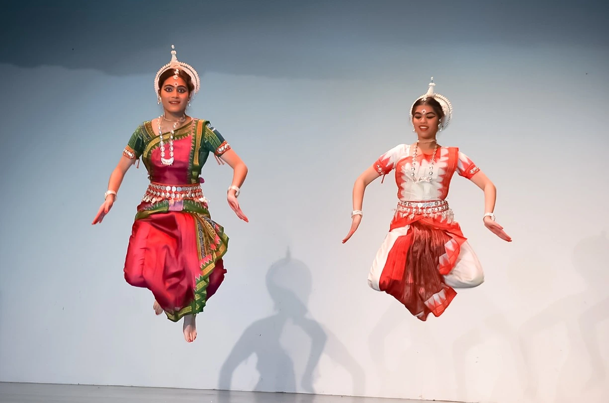 Two Odissi dancers perform a synchronized mid-air jump in traditional attire